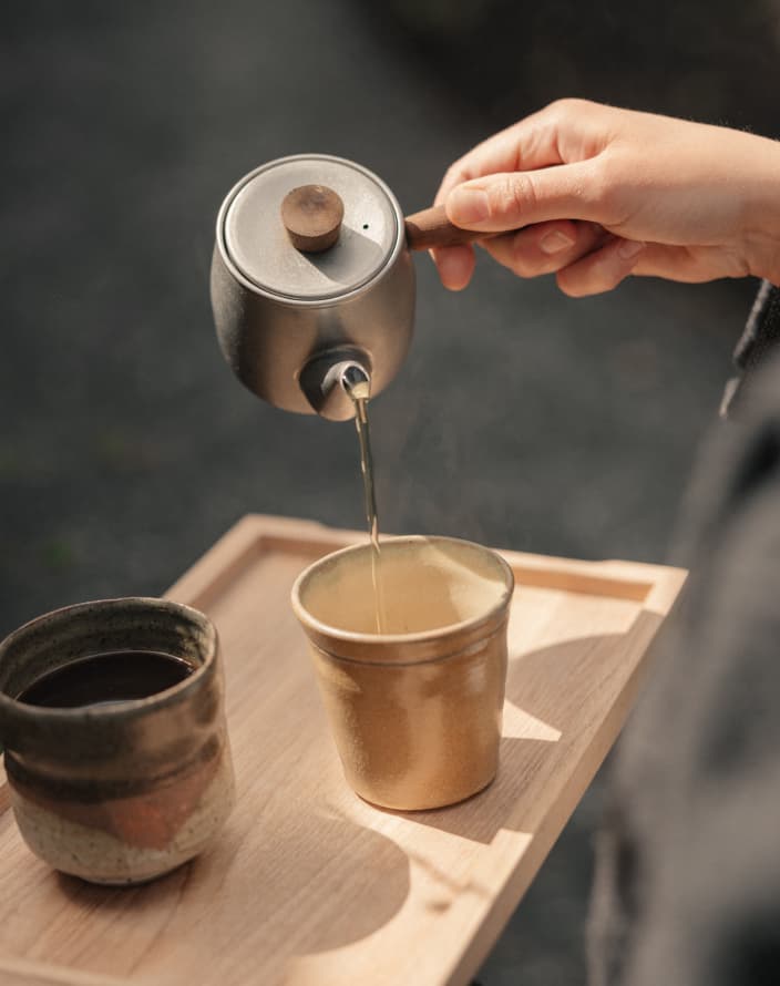 A hand pouring tea from a metal pot into a small ceramic cup